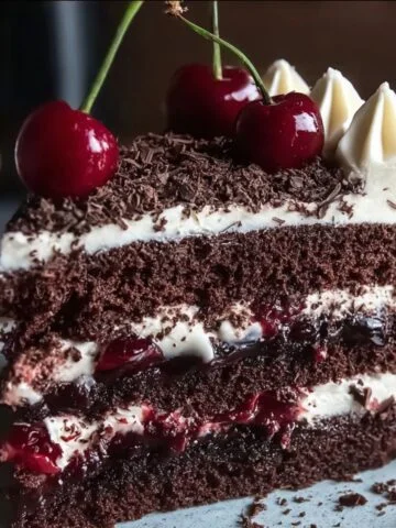 A slice of homemade Black Forest cake with chocolate layers, whipped cream, cherry filling, dark chocolate shavings, and fresh cherries on top.