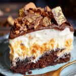 Homemade Butterfinger cake slice with a chocolate base, creamy filling, candy pieces, and chocolate ganache topped with chopped Butterfinger and gold leaf, served on a blue plate with a gold fork.