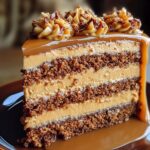 A slice of homemade caramel cake with four moist layers, rich caramel frosting, and a glossy caramel drizzle, topped with piped frosting and chopped nuts on a brown plate.