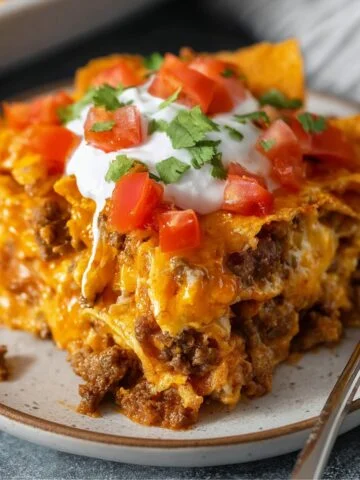 Homemade Dorito casserole slice topped with sour cream, diced tomatoes, and cilantro, served on a plate with melted cheese and crunchy chips visible.