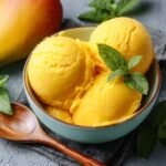 Three scoops of bright yellow mango sorbet in a teal bowl, topped with fresh mint, next to a wooden spoon and a ripe mango.