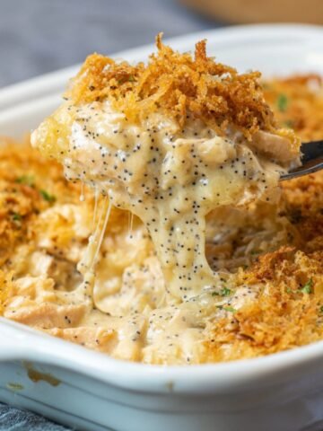 A fork lifts a creamy, cheesy scoop of homemade poppy seed chicken casserole from a white baking dish, showing tender chicken, black poppy seeds, and a golden, crispy topping.