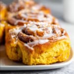 Homemade pumpkin cinnamon rolls topped with cream cheese glaze, showing golden-orange dough swirls and gooey spiced filling on a white plate.