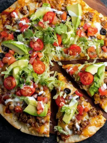 Homemade taco pizza topped with ground beef, cheese, lettuce, tomatoes, avocado, and sour cream on a wooden board.