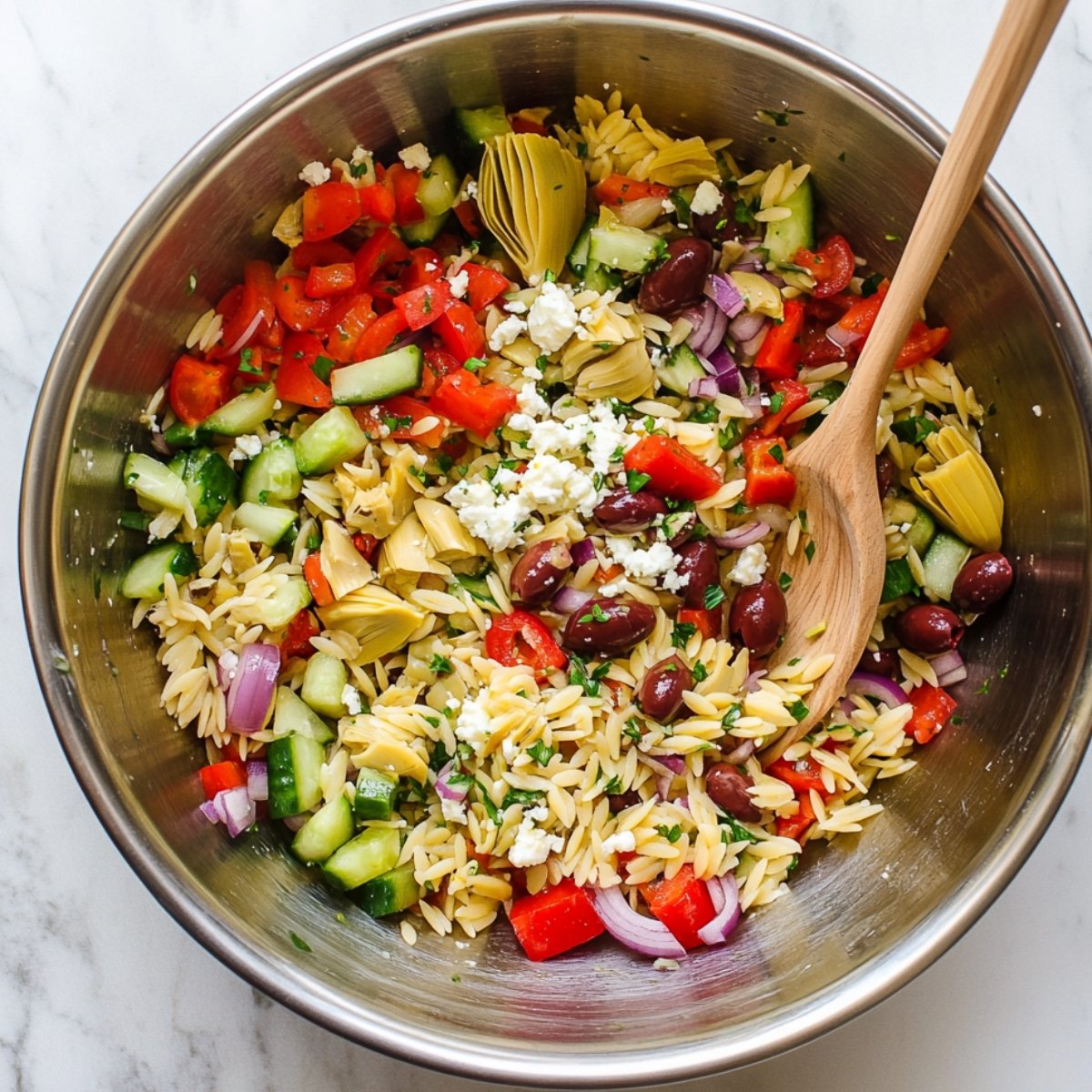Greek orzo salad in a metal bowl with cucumber, red pepper, olives, artichoke hearts, red onion, feta, and herbs, mixed with a wooden spoon.