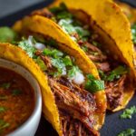 Three crispy homemade birria tacos filled with shredded beef, topped with fresh cilantro and diced onion, served with lime wedges and a bowl of rich consommé for dipping.