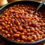 Golden, homemade baked beans in a round cast-iron Dutch oven, topped with crispy bacon and garnished with fresh thyme, showing a caramelized surface from slow oven baking.