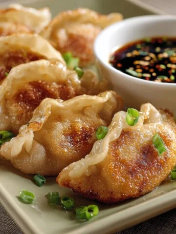 Homemade fried dumplings with crispy golden bottoms and pleated tops, garnished with green onions, served with soy dipping sauce in a white bowl.
