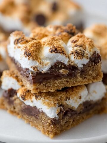 Homemade s’mores bars on a white plate, showing layers of golden graham cracker crust, melted chocolate, and toasted marshmallows with lightly charred tops. Blurred bars in the background complete the cozy, homemade dessert look.