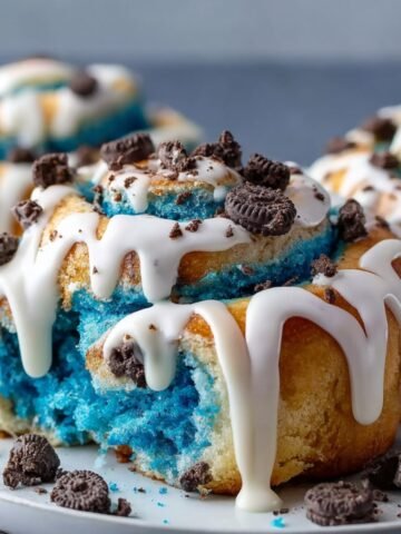 Homemade Cookie Monster cinnamon rolls with blue swirled dough, white icing drizzle, Oreo crumbles, and golden edges.