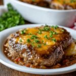 Homemade Amish hamburger steak bake topped with melted cheddar cheese and rich brown gravy, garnished with parsley and served over mashed potatoes on a white plate.