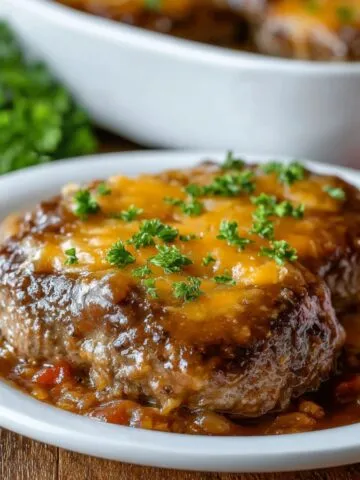 Homemade Amish hamburger steak bake topped with melted cheddar cheese and rich brown gravy, garnished with parsley and served over mashed potatoes on a white plate.