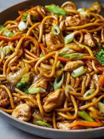 Chicken Lo Mein Recipe in a skillet with glossy noodles, tender chicken pieces, broccoli florets, carrots, onions, and scallions tossed in a rich soy-based sauce - perfectly caramelized and ready to serve, showing real home-cooked texture and freshness.