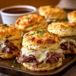 French Dip Biscuits filled with roast beef and melted cheese, topped with herbs and served with au jus on a wooden board in warm natural light.