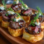 Beef Wellington Bites topped with seared medium-rare beef, sautéed mushrooms, blue cheese crumbles, and rosemary on golden puff-pastry rounds.