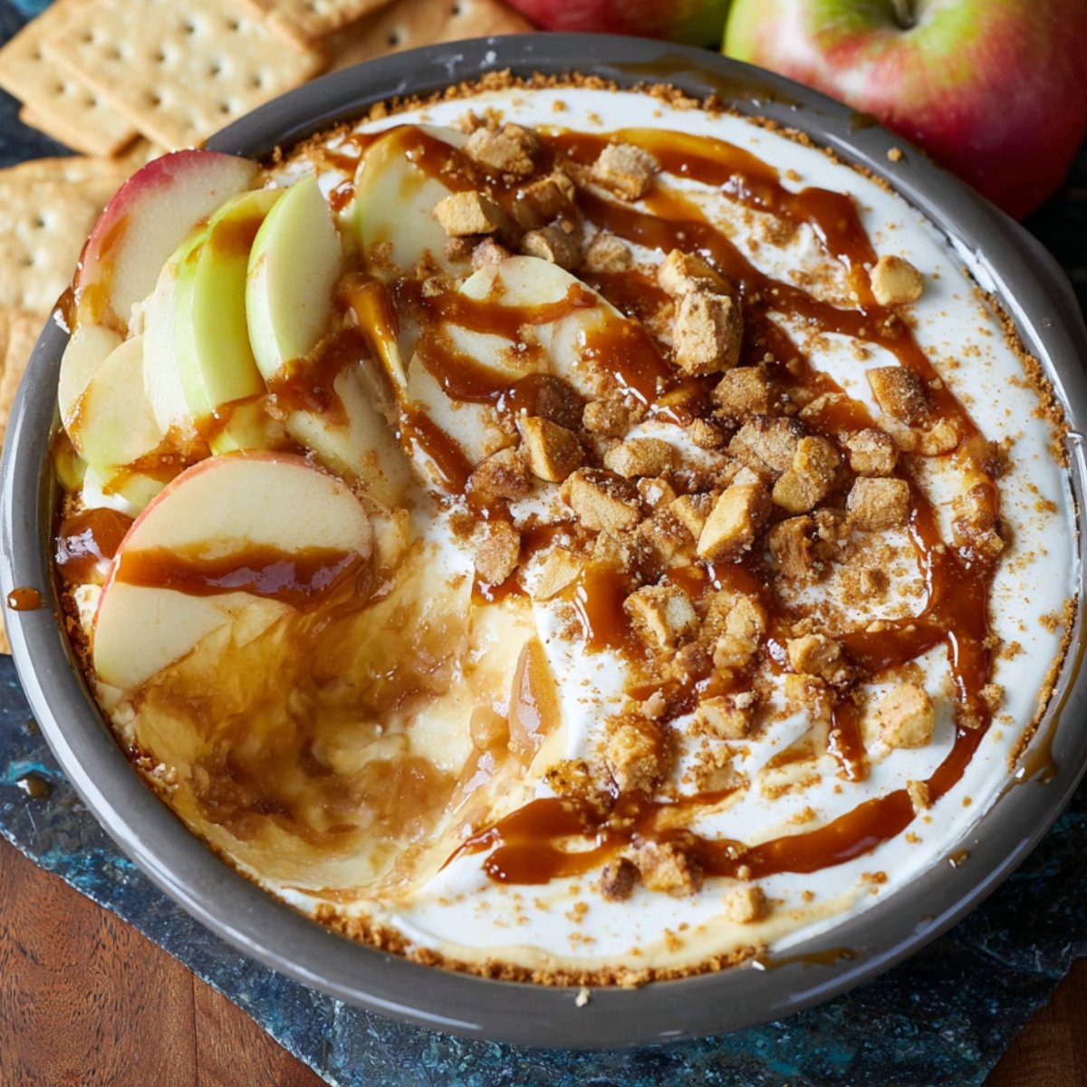 Homemade caramel apple cheesecake dip topped with sliced apples, caramel drizzle, and crumbled graham crackers in a round dish, with fresh apples and crackers in the background.