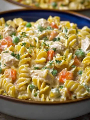 A bowl of creamy homemade chicken pot pie pasta with rotini noodles, tender chicken pieces, carrots, and peas, coated in a rich, savory sauce and topped with fresh parsley.