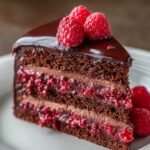 Homemade chocolate raspberry layer cake slice with glossy chocolate ganache, rich cocoa sponge, and bright raspberry filling topped with fresh raspberries on a white plate.