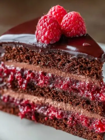 Homemade chocolate raspberry layer cake slice with glossy chocolate ganache, rich cocoa sponge, and bright raspberry filling topped with fresh raspberries on a white plate.
