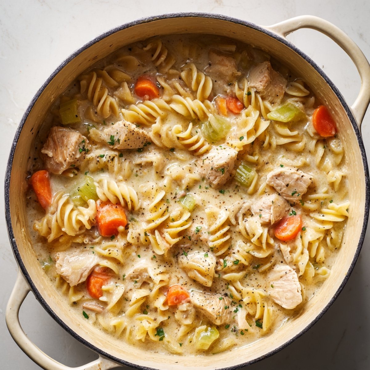 Homemade chicken pot pie pasta in a cream-colored Dutch oven, filled with tender rotini noodles, chunks of chicken, sliced carrots, and celery in a rich, creamy sauce, seasoned with black pepper and parsley.