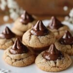 Homemade peanut butter blossom cookies topped with soft, glossy chocolate kisses, lightly coated in sugar and arranged on a plate with a warm, cozy background.