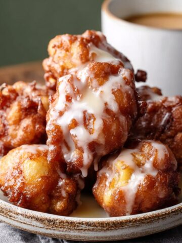 Homemade Apple Fritters drizzled with glaze, golden brown and crisp, served on a rustic ceramic dish beside a cup of coffee.