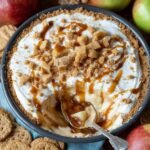 Homemade caramel apple cheesecake dip topped with caramel drizzle, crumbly brown sugar streusel, and diced apples in a round dish, surrounded by apples and graham crackers.