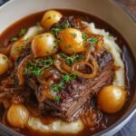 Homemade French onion pot roast served over mashed potatoes, topped with caramelized onions, pearl onions, and fresh thyme in rich brown gravy, looking tender, glossy, and comforting.