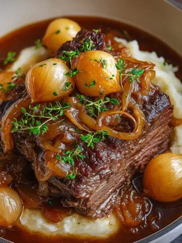 Homemade French onion pot roast served over mashed potatoes, topped with caramelized onions, pearl onions, and fresh thyme in rich brown gravy, looking tender, glossy, and comforting.