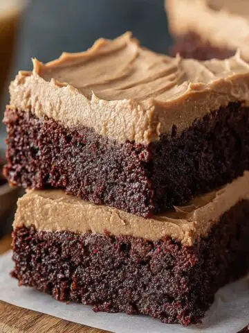 Homemade Frosted Coffee Brownies stacked on parchment paper, showing a dense, fudgy chocolate base and thick, creamy coffee buttercream frosting on top, with soft texture and natural crumbs visible, set on a wooden board for a cozy, real kitchen look.