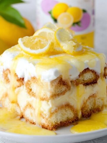 Homemade Lemon Tiramisu with layers of ladyfingers, lemon cream, whipped topping, and lemon curd dripping down the sides, topped with fresh lemon slices on a white plate.