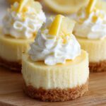 Homemade mini lemon cheesecakes with a golden graham cracker crust, topped with whipped cream, lemon zest, and a fresh lemon slice, displayed on a wooden board.