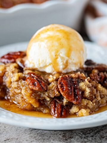 Homemade Pecan Pie Cobbler topped with a scoop of melting vanilla ice cream, featuring caramelized pecans and a gooey brown sugar filling on a white dessert plate.