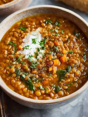 A warm bowl of homemade lentil soup topped with fresh herbs and a swirl of cream, served with bread on the side.
