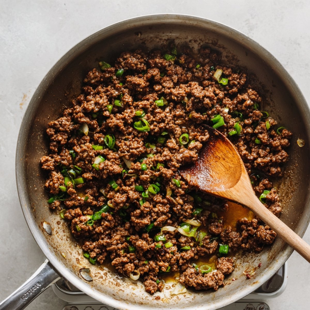 Homemade Korean ground beef cooking in a skillet with chopped green onions and sauce, stirred with a wooden spoon.