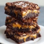 Stack of homemade Outrageous Snickers Brownies with rich chocolate layers, gooey caramel filling, and a glossy drizzle on top, sprinkled with flaky sea salt, served on a white plate for a delicious, real, and freshly baked look.