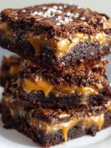 Stack of homemade Outrageous Snickers Brownies with rich chocolate layers, gooey caramel filling, and a glossy drizzle on top, sprinkled with flaky sea salt, served on a white plate for a delicious, real, and freshly baked look.