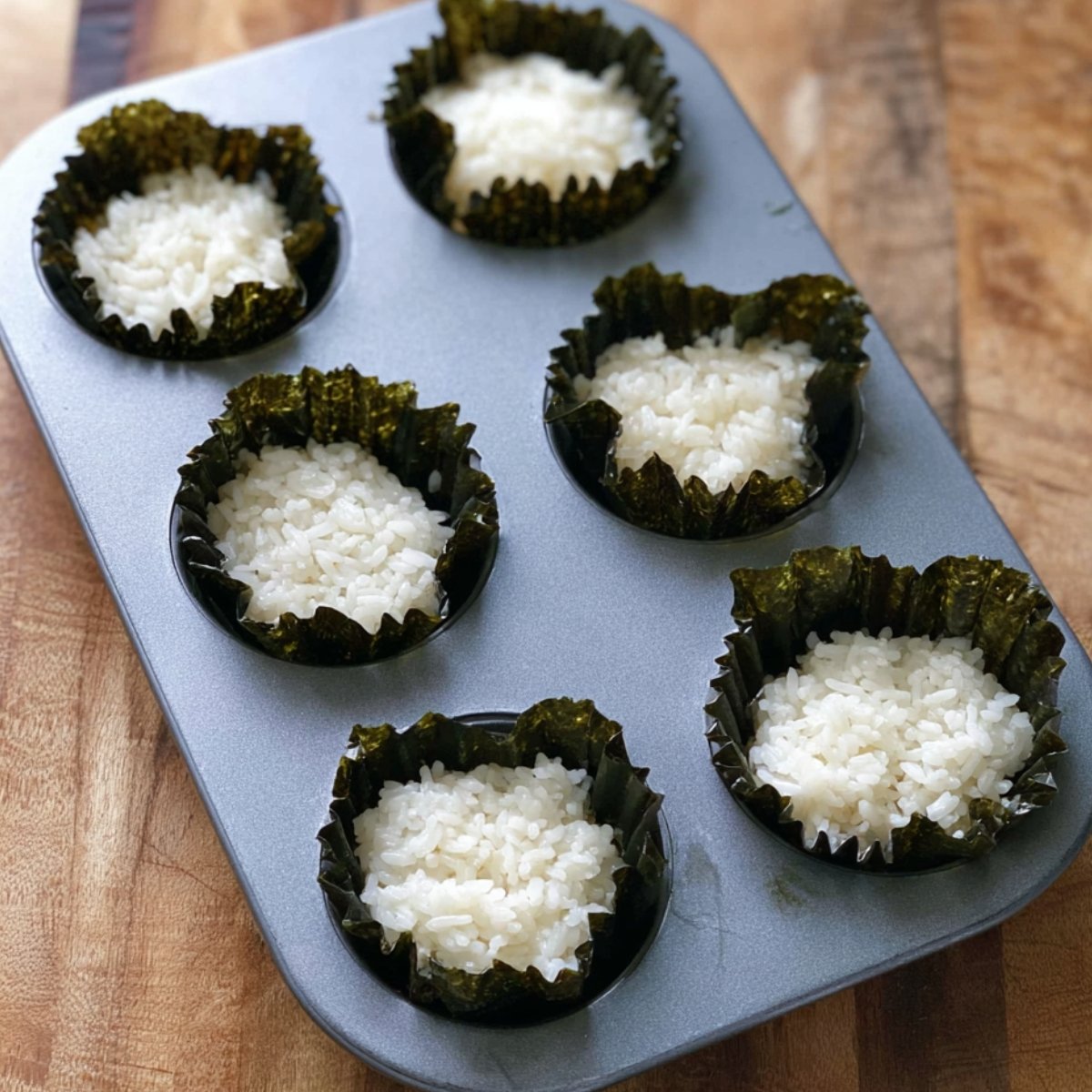 Place one nori square into each muffin cup. Add 2 tablespoons of sushi rice on top of each nori sheet, then use your fingers or the back of a spoon to press the rice down gently. You want it to stick to the nori and form a little base.