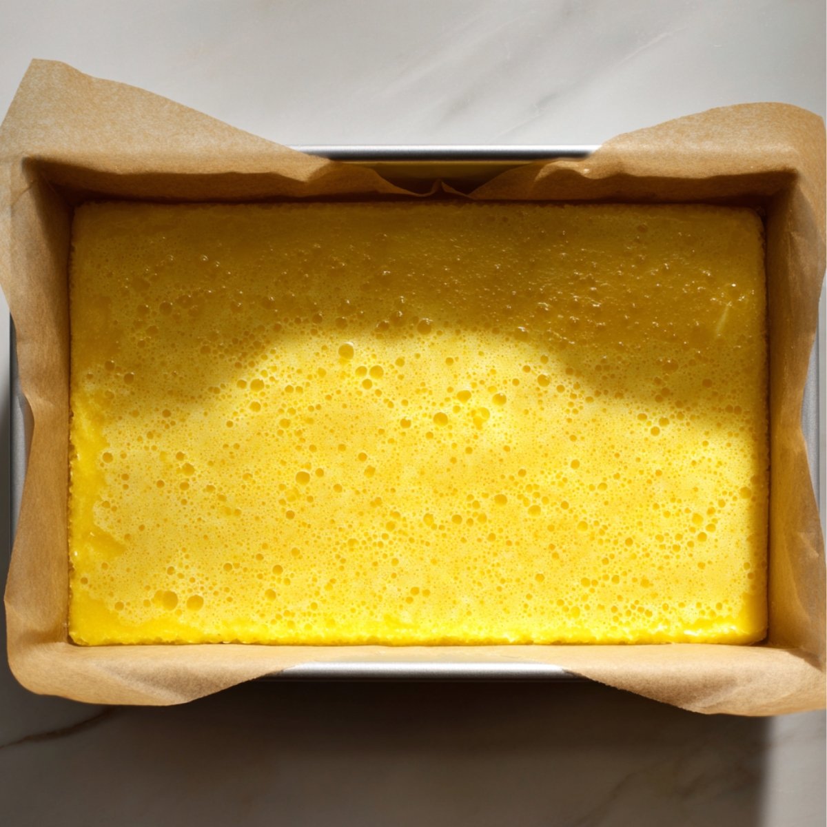 Freshly baked homemade lemon bars in a parchment-lined pan, with a bright yellow, slightly bubbly surface under natural sunlight.
