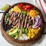 A homemade Cilantro Lime Steak Bowls filled with sliced grilled steak, rice, black beans, avocado, corn, tomatoes, red onion, jalapeños, shredded cheese, and a creamy drizzle, all arranged neatly in a rustic wooden bowl.