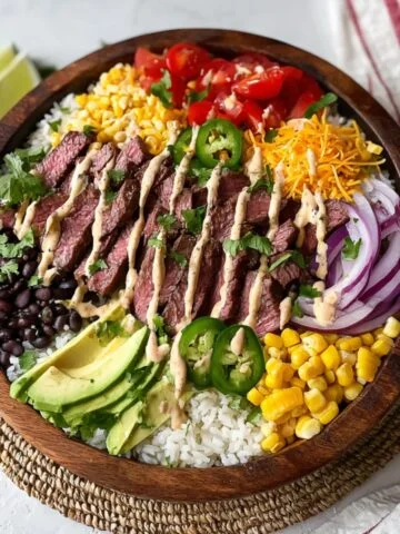 A homemade Cilantro Lime Steak Bowls filled with sliced grilled steak, rice, black beans, avocado, corn, tomatoes, red onion, jalapeños, shredded cheese, and a creamy drizzle, all arranged neatly in a rustic wooden bowl.
