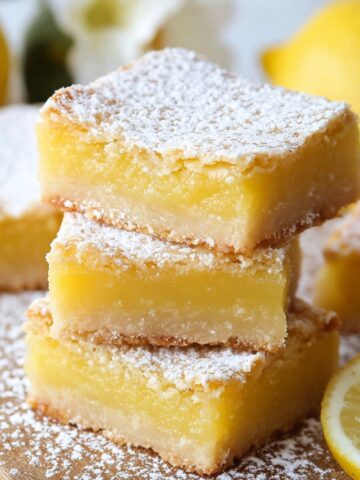 Homemade Lemon Bars dusted with powdered sugar, sitting on a wooden board with fresh lemons in the background.