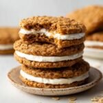 Homemade Oatmeal Cream Pies with one showing a bite taken out, revealing the creamy filling between the oatmeal cookies. The cookies are golden brown and placed on a simple ceramic plate.