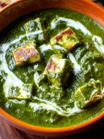 A bowl of creamy Palak Paneer with paneer cubes in a vibrant spinach gravy, topped with a swirl of cream, served with naan.