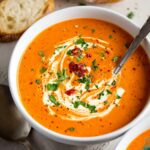 Homemade Roasted Red Pepper Gouda Soup, garnished with a swirl of cream, fresh parsley, and sun-dried tomatoes. A spoon is placed in the bowl, and a piece of bread is visible beside it.
