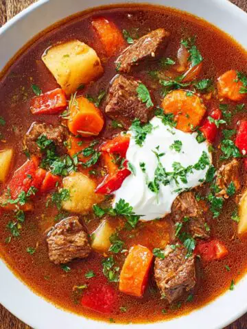 A hearty bowl of homemade Hungarian goulash filled with tender beef, potatoes, carrots, and red peppers, topped with a dollop of sour cream and fresh parsley.