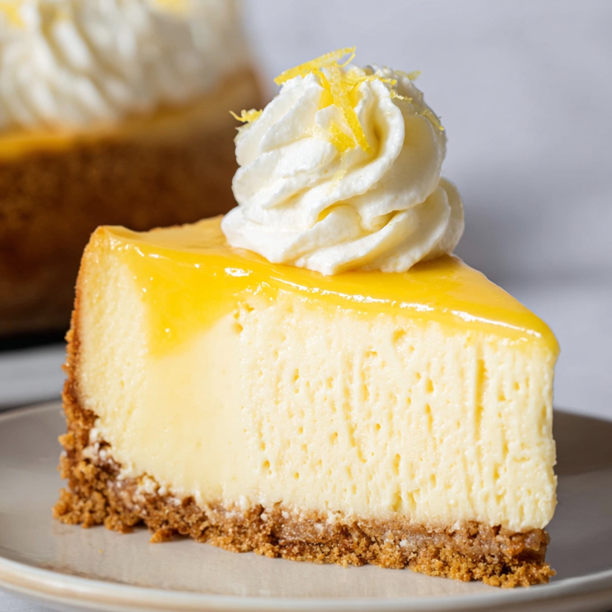 A slice of lemon cheesecake with a golden curd topping, whipped cream, and a graham cracker crust.
