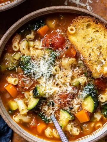 A close-up of a bowl of minestrone soup with pasta, vegetables, and grated cheese, served with a slice of toasted bread.