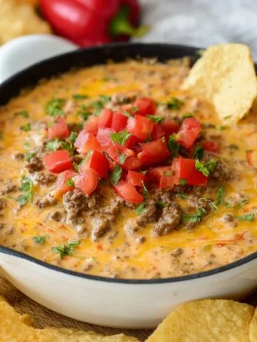 Rotel Dip with Ground Beef, topped with fresh chopped tomatoes and green onions, served in a white bowl surrounded by crispy tortilla chips. The dip has a creamy texture with melted cheese and visible beef.