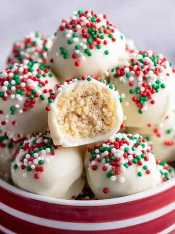 Sugar cookie truffles covered in white chocolate and colorful holiday sprinkles, with one truffle broken open to show the crumbly filling inside.